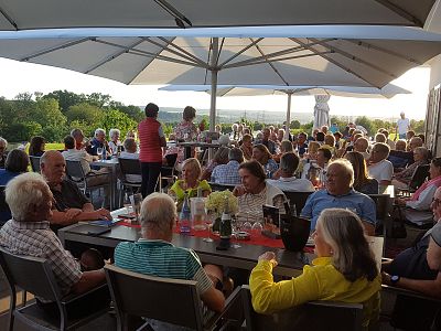 A patio filled with people gathered at tables under large umbrellas. The scene is bright and social, with greenery visible in the background.