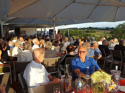 A large outdoor gathering with people seated under umbrellas, enjoying the sunset view from a terrace overlooking a scenic landscape.