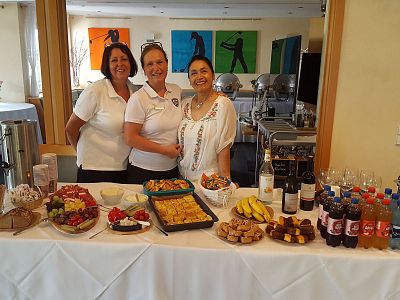 Three people stand behind a table with a variety of food and drinks, including fruits, pastries, and beverages, in a room with colorful paintings.