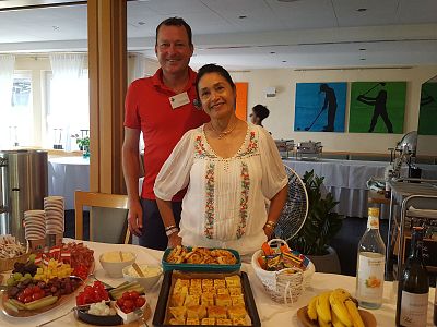 Two people stand behind a table with a variety of food items, including fruits and snacks, smiling at the camera. The setting appears to be a warm indoor event.