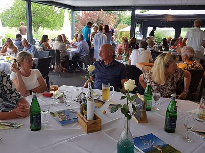 A group of people dining outdoors under a canopy, surrounded by greenery, enjoying food and drinks at elegantly set tables.