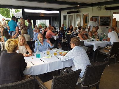 A large group of people dining at an outdoor event, seated at long tables with white tablecloths. The atmosphere is lively and social, with groups chatting.