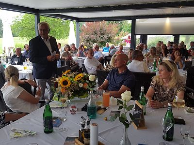 People seated at round tables in a bright room, listening to a speaker, with flowers and drinks on the tables.
