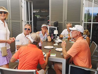 A group of six people sit and stand around an outdoor table, smiling and talking, dressed in casual summer attire, with meals and drinks on the table.