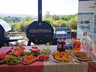 A table with a variety of snacks, fruits, and drinks is set up outdoors with BBQ grills and a scenic view in the background.