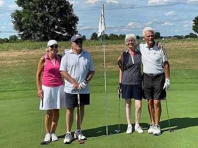 Four people stand on a golf course green with a flag in the background, all smiling and holding golf clubs, under a bright blue sky with scattered clouds.