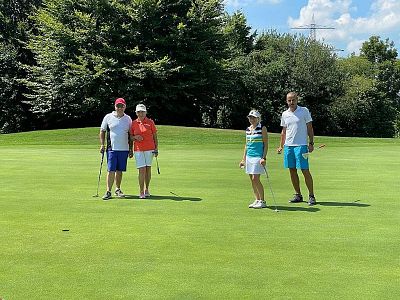 Four people on a golf course, standing on the green with clubs. They are in casual attire, surrounded by trees and a bright blue sky.
