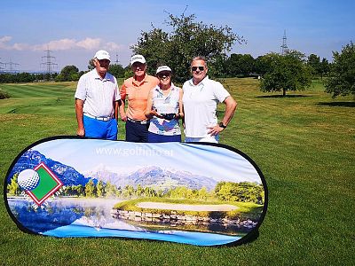 Four people posed on a golf course next to a banner with a scenic landscape. Trees and power lines in the background, indicating a sunny day.