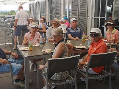 A group of people seated at a long table outside, enjoying a meal. They wear hats and sunglasses, with a clear sky and open umbrellas in the background.