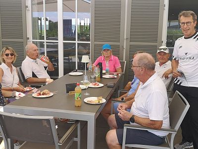 A group of people sitting at an outdoor table, eating and drinking. They are casually dressed and appear to be enjoying a meal together in a sunny setting.