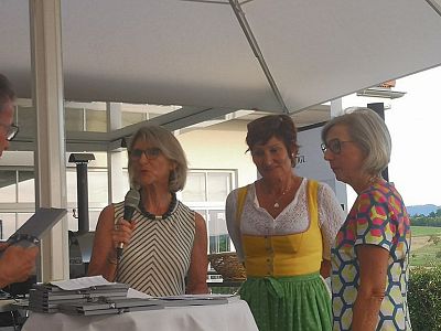 Three women and one man standing under a canopy, one speaking into a microphone, others listening. Outdoor event with greenery in the background.