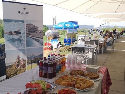 A table with pastries and drinks at an outdoor event. People sit at tables under umbrellas. A banner and a scenic background are visible.
