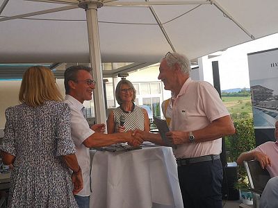 Group of people shaking hands and conversing at an outdoor event under a large umbrella.