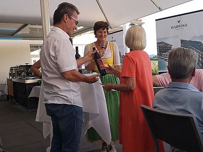 A group of people socializing at an outdoor event. A man hands a bottle to a seated woman, while another woman stands smiling. Banners and tables are visible.