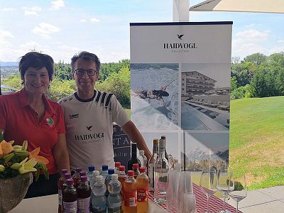 Two people stand behind a table with drinks and a promotional banner. The background shows a green landscape. Bottled beverages are displayed on the table.