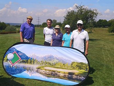 Five people standing on a golf course behind a banner with a scenic image. They are dressed casually, with golf equipment visible. Trees and sky in the background.