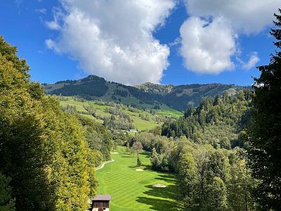 A green valley with a golf course, surrounded by mountains and trees under a blue sky with white clouds in Switzerland.