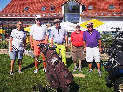 Five men in golf attire stand together on a sunny golf course. A clubhouse with a red roof and yellow umbrellas is visible in the background.