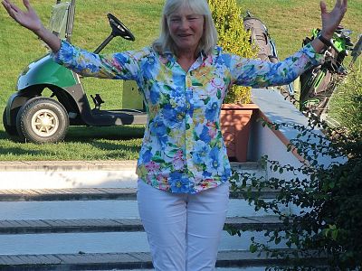 A woman in a floral shirt and white pants stands with arms open on steps. A golf cart and greenery in the background suggest an outdoor setting.
