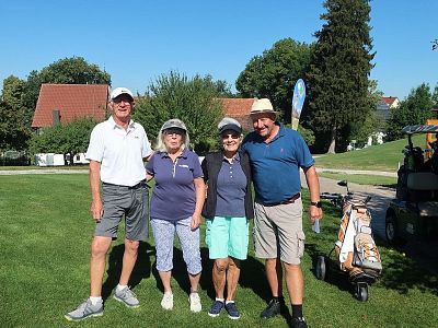 Four smiling individuals in golf attire stand on a green field with trees and a golf cart nearby, enjoying a sunny day.
