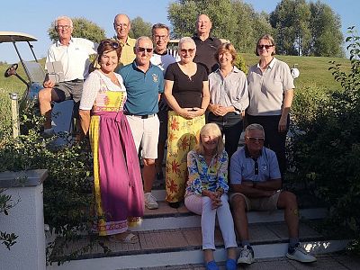 A group of people, some seated, others standing on stone steps outdoors. One wears traditional attire. Greenery and a golf cart are in the background. Sunny day.