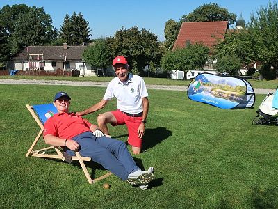 Two men on a golf course, one sitting in a deck chair with a cap, the other crouching beside him smiling, both in sporty clothes, sunny day.
