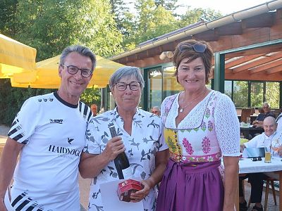 Three people pose outdoors at a café; one holds a wine bottle, surrounded by greenery, under a sunny sky with yellow umbrellas.