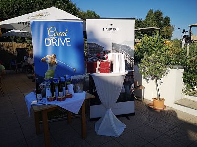Outdoor setting with golf-themed banners, a table displaying wine bottles and a gift box, trees and blue sky in the background.