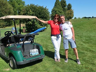 Two people stand by a golf cart on a green golf course. They appear cheerful, with trees and blue sky in the background, suggesting a pleasant outdoor setting.