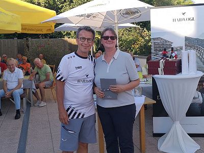Two people posing for a photo at an outdoor event, with tables and umbrellas in the background. They are smiling and dressed casually.