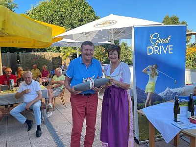 A man in a blue shirt and red pants, with a woman in a traditional dress, holding a prize at an outdoor event. People sit at tables under umbrellas.