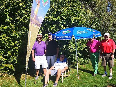 A group of people, one seated under an umbrella, pose casually outdoors in a sunny setting by trees, suggesting a relaxed atmosphere at a golf event.
