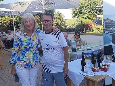 Two people smiling at a wine tasting event outdoors. Tables with wine bottles and shade umbrellas are visible. Others are seated in the background.