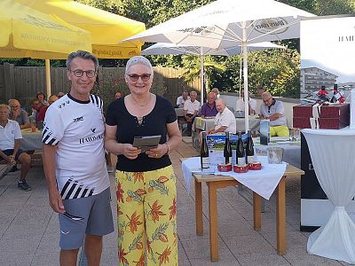 People gather outdoors under umbrellas, with wine bottles and gifts on tables. The setting is casual, suggesting a social event in sunny weather.