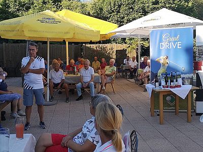 People seated at tables outdoors, listening to a speaker. Yellow umbrellas and a promotional banner are in the background, suggesting a social gathering.