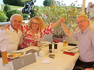 Three people sit at an outdoor restaurant table with drinks and food. They smile and hold hands, celebrating a joyful moment under a canopy with greenery.