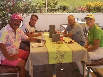 Four people seated at an outdoor table, surrounded by greenery, sharing a meal. They're dressed casually, with a mix of colorful and neutral clothing.