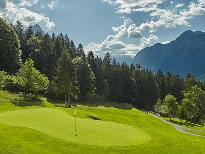 A lush green golf course with a mountainous backdrop under a cloudy sky, surrounded by dense forests, depicting a serene and scenic alpine landscape.