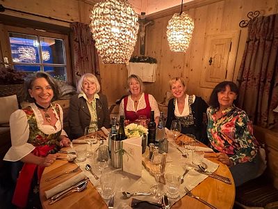 Five women in traditional attire sit around a wooden dining table with elegant place settings in a warmly lit room.