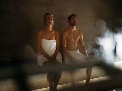 A man and woman in towels sit in a dimly lit steam room, highlighting a calm and relaxed spa atmosphere.
