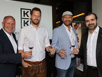 Vier Männer in Anzügen stehen nebeneinander und halten Weingläser. Sie lächeln in die Kamera. Im Hintergrund ist ein modernes Interieur sichtbar.