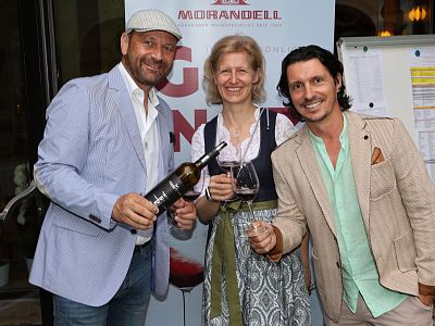 Drei Personen stehen nebeneinander, zwei Männer und eine Frau. Sie halten Weingläser und lächeln in die Kamera. Hintergrund zeigt ein Plakat und einen Flipchart.