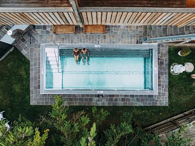 Aerial view of a modern rectangular pool with stone tiles and greenery, where two people swim casually, creating a calm, relaxing atmosphere.
