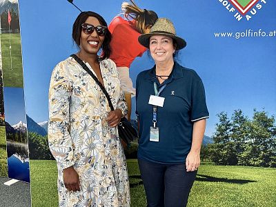 Two smiling people at an event, one in a floral dress and sunglasses, the other in a polo shirt with a hat, standing in front of a scenic backdrop.