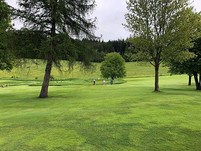 A lush green golf course with rolling hills and trees. Two people play golf in the distance under a cloudy sky.