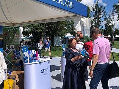 People engage at a booth with sustainable living materials; trees and a lawn in the background, suggesting an outdoor fair setting.
