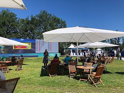 People seated on wooden chairs under umbrellas at an outdoor event. Bright day with a large screen in the background and green grass.