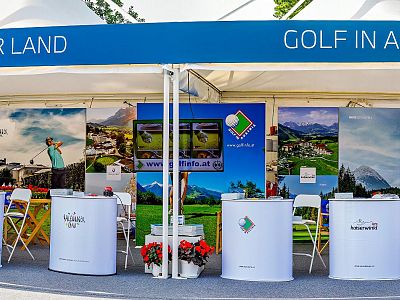 Booth featuring golf destinations in Austria with banners, chairs, and brochures under a blue and white canopy at a travel fair.