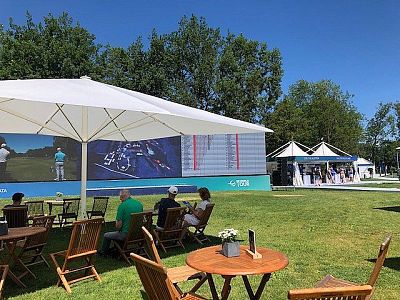 Outdoor seating area with wooden tables and chairs on grass, under umbrellas. A large screen shows a golf game; tents are in the background, trees around.