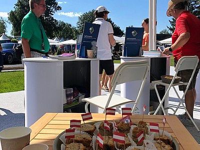 Small outdoor event with people at white booths under a sunny sky, displaying brochures. A plate with small flags and snacks is on a table in the foreground.